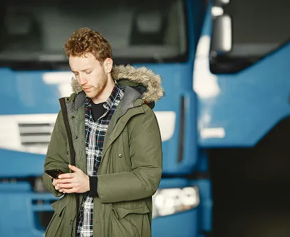 Young man wearing a green parka jacket and plaid shirt, looking at his smartphone with blue trucks in the background.