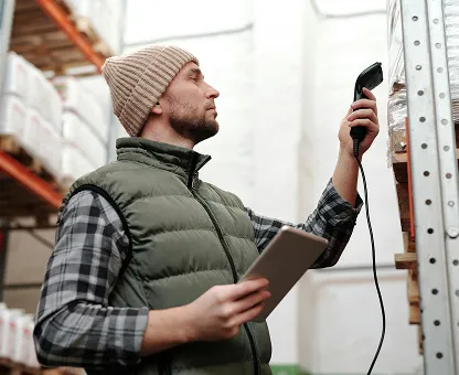 Man in a beanie and vest scanning barcodes on shelves in a warehouse while holding a tablet.