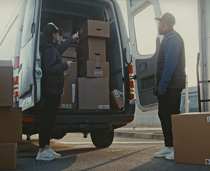 Two delivery workers unloading stacked cardboard boxes from the back of a white van in an outdoor setting.