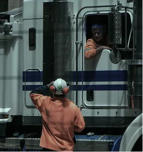 One trucker in an orange shirt and headphones stands outside a large white truck talking to another trucker leaning out the driver's window.