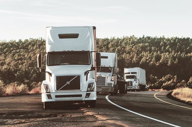 Three white semi-trucks parked in a line on a curved road with forested hills in the background.