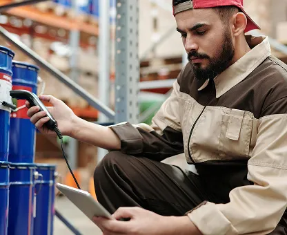 Worker in brown and beige uniform measuring blue barrels with a handheld device in a warehouse.