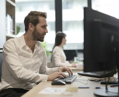 A man in a white shirt typing on a keyboard at a desk with a large monitor in an office setting.