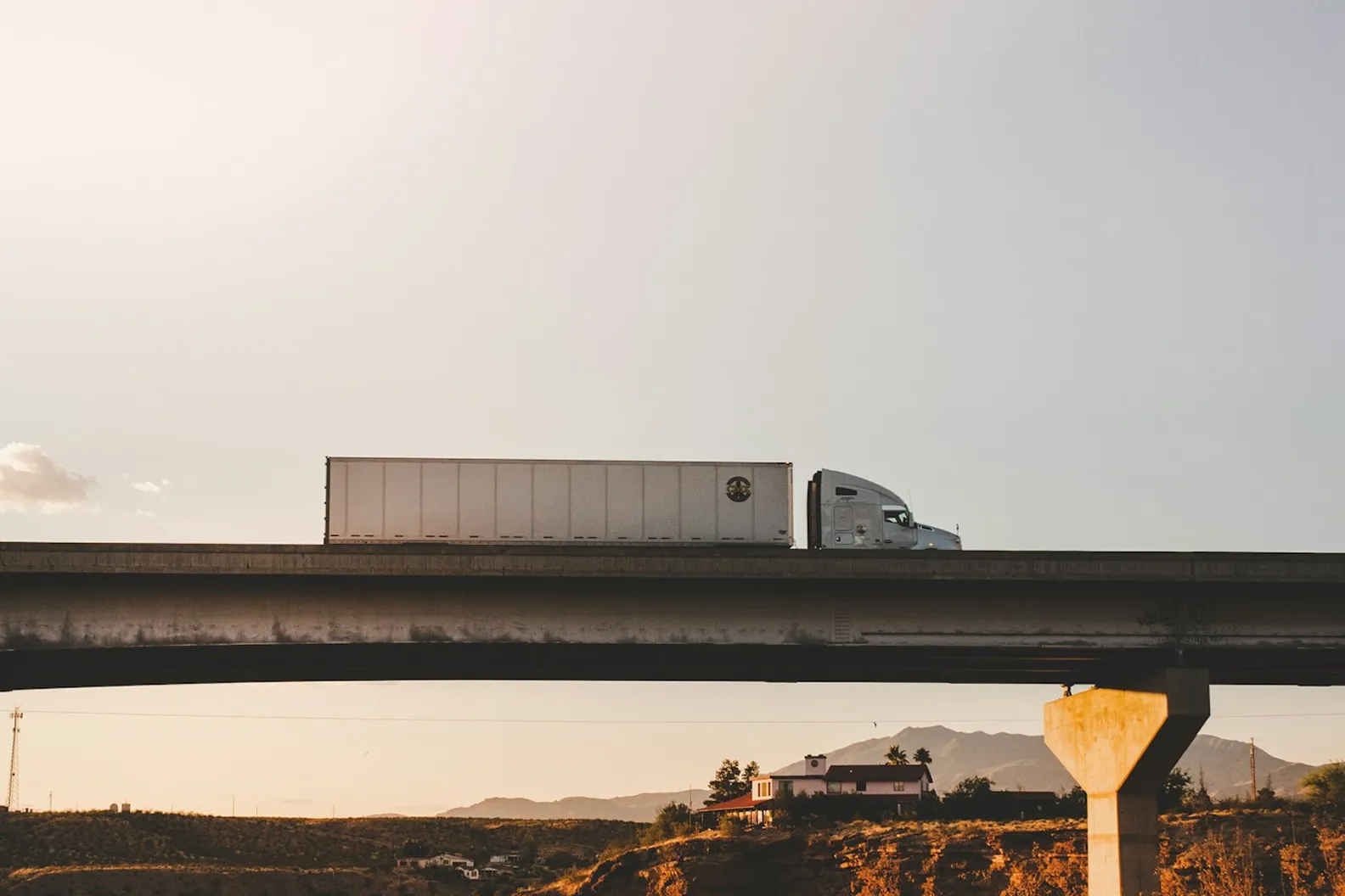 White semi-truck driving on a highway overpass above a desert landscape with a house and mountains in the background during sunset.