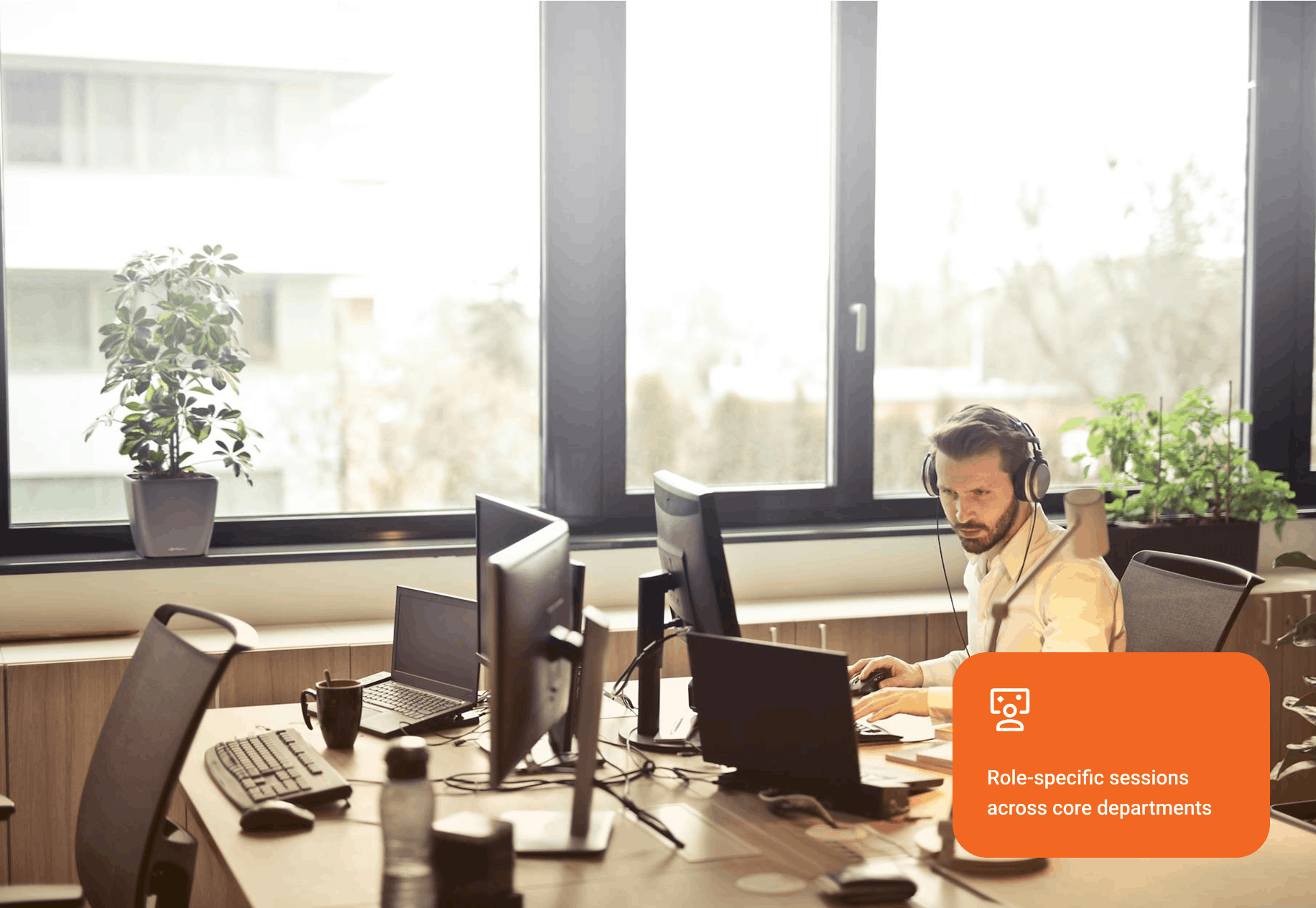 Man wearing headphones working on a laptop at a desk with multiple monitors in a bright office with large windows and plants.