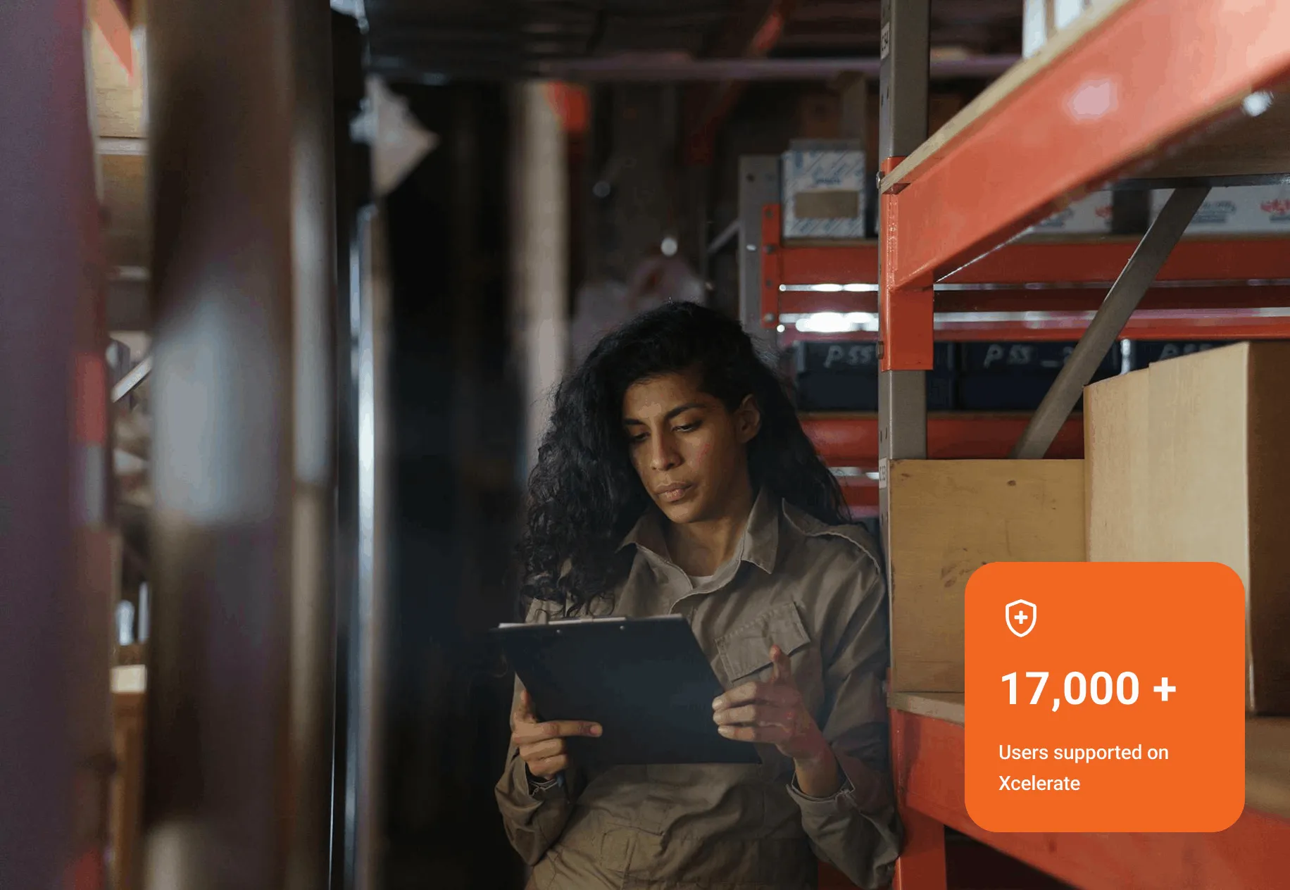 Worker in a uniform checking a clipboard while sitting next to orange metal shelves with boxes in a warehouse.
