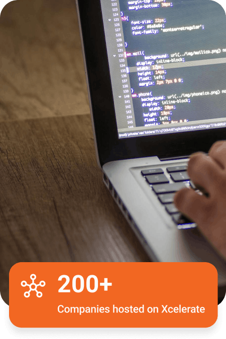 Partial view of a laptop displaying code on screen with a hand on the keyboard, and an orange overlay stating '200+ Companies hosted on Xcelerate'.