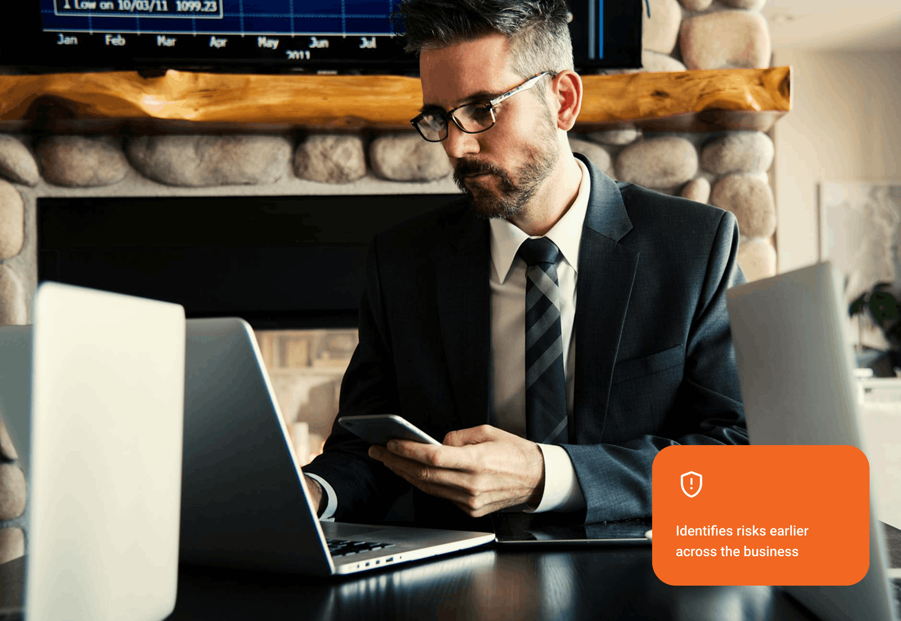 Man in a suit and glasses working on a laptop and holding a smartphone with a financial chart in the background.