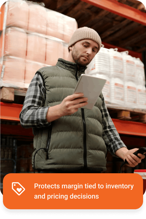 Man in a beanie and green puffer vest holding a tablet and scanning inventory in a warehouse.