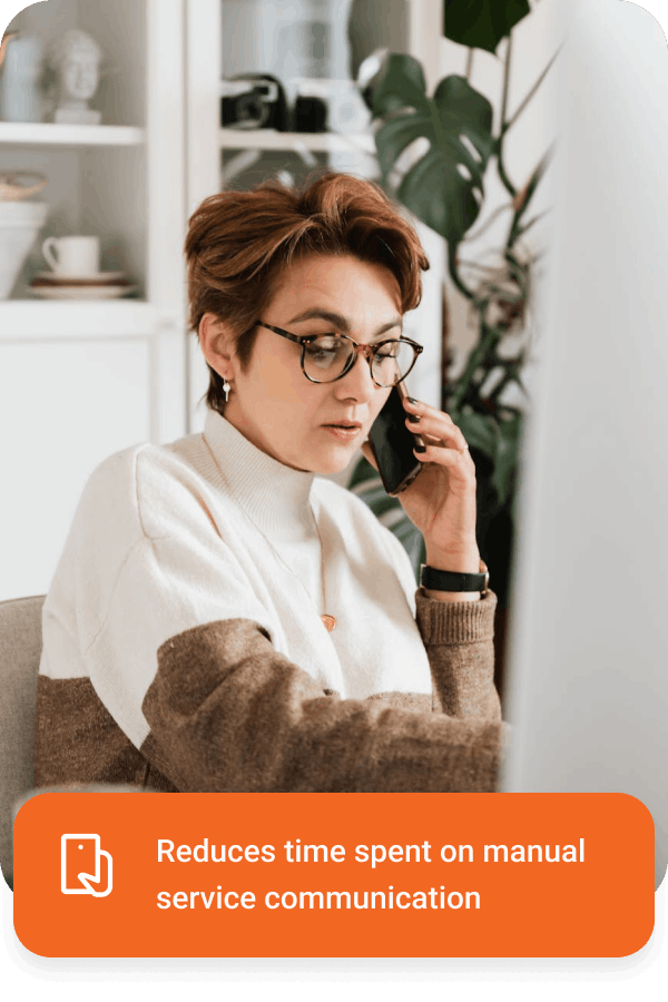 Woman wearing glasses talking on a smartphone while working at her desk with a plant in the background.