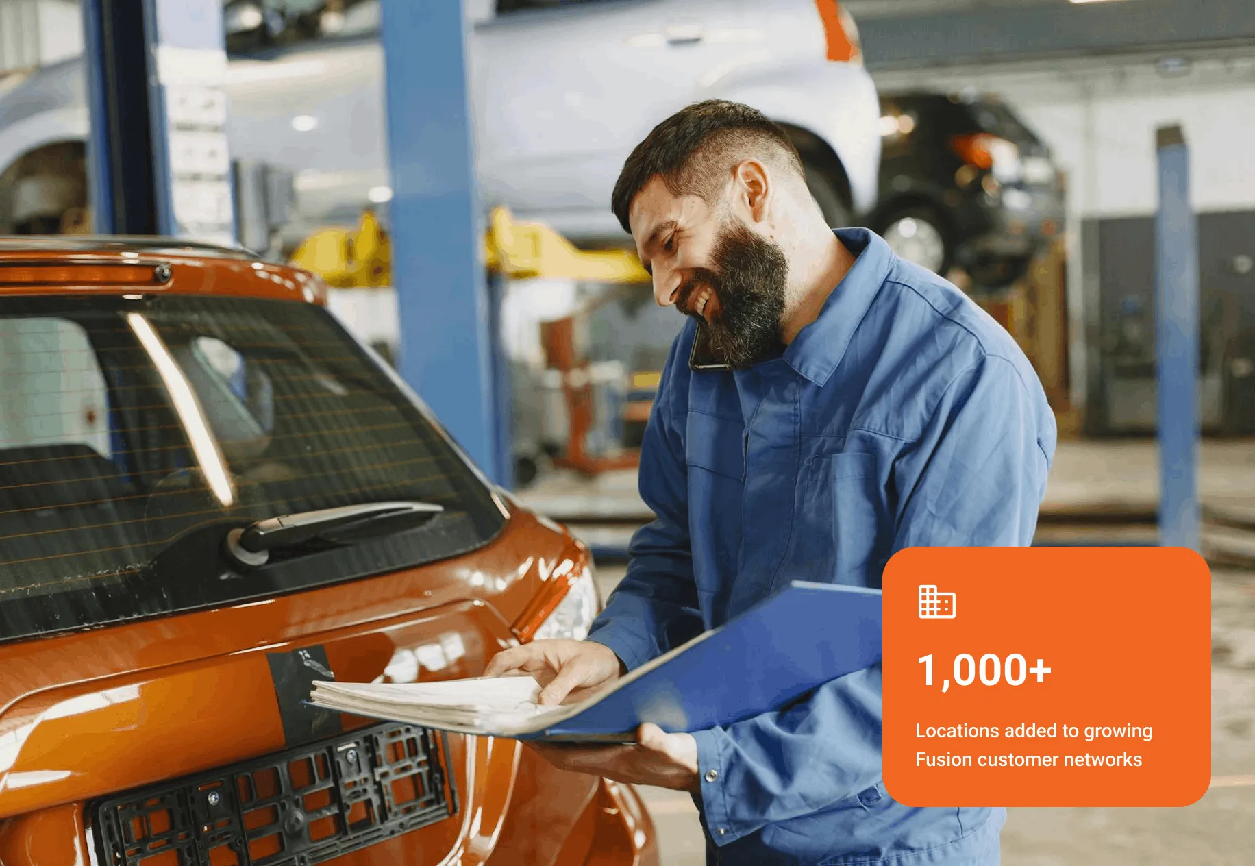 Mechanic in blue uniform holding a clipboard and talking on phone next to an orange car in a garage.