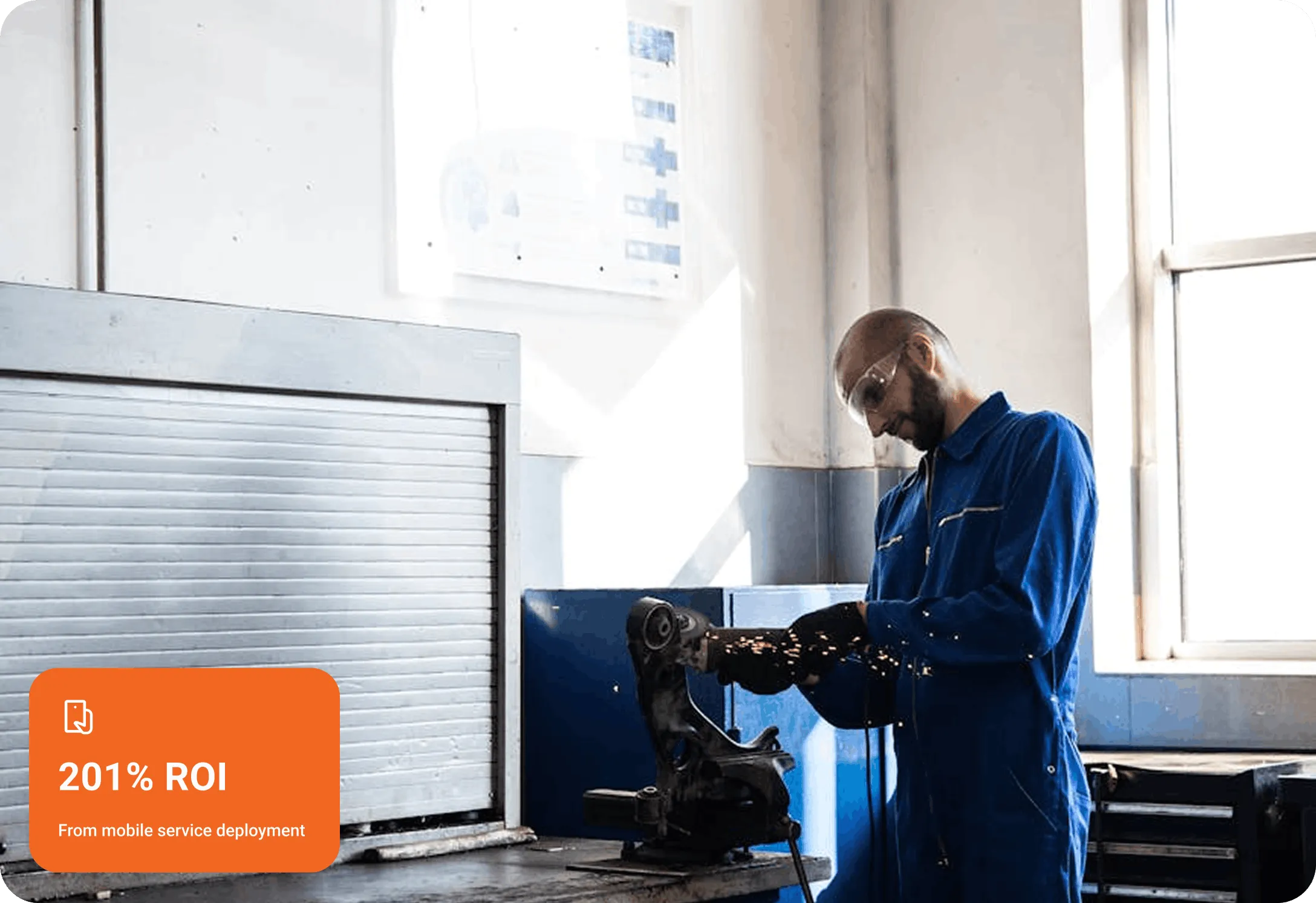Worker in blue coveralls and safety glasses using a grinding tool on a metal part in a workshop with sparks flying.