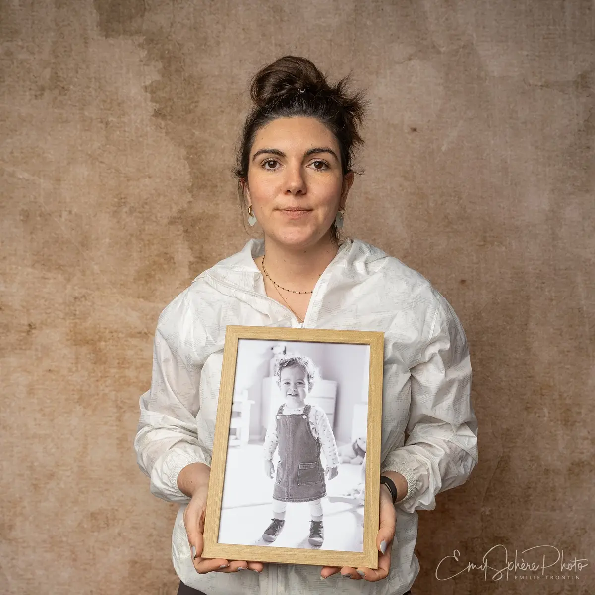 Photo pour le portrait de Laura, maman d'Emie. Elle pose seule devant un fond sable. Elle porte dans les mains, un portait d'Emie dans un cadre en bois.
