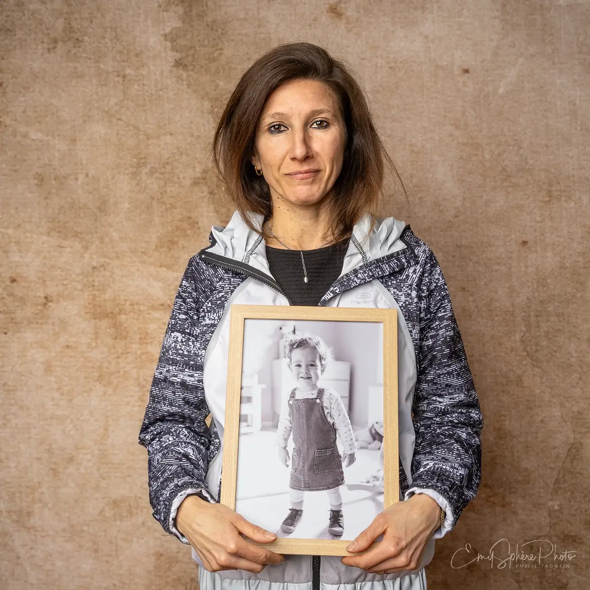 Photo pour le portrait d'Aurelia, maman d'Emie. Elle pose seule devant un fond sable. Elle porte dans les mains, un portait d'Emie dans un cadre en bois.