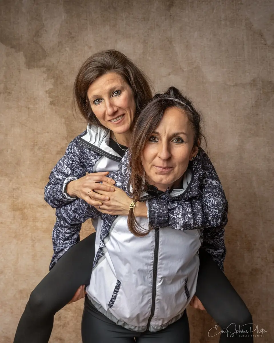 Photo pour la présentation du duo sportif, Aurelia (maman d'Emie) et Maryline (marraine d'Emie). Elles posent à deux devant un fond sable. Habillées en tenue de sport. Maryline porte Aurelia sur ses épaules, symbole de leur force, de leur amitié et de l'entraide entre elles.