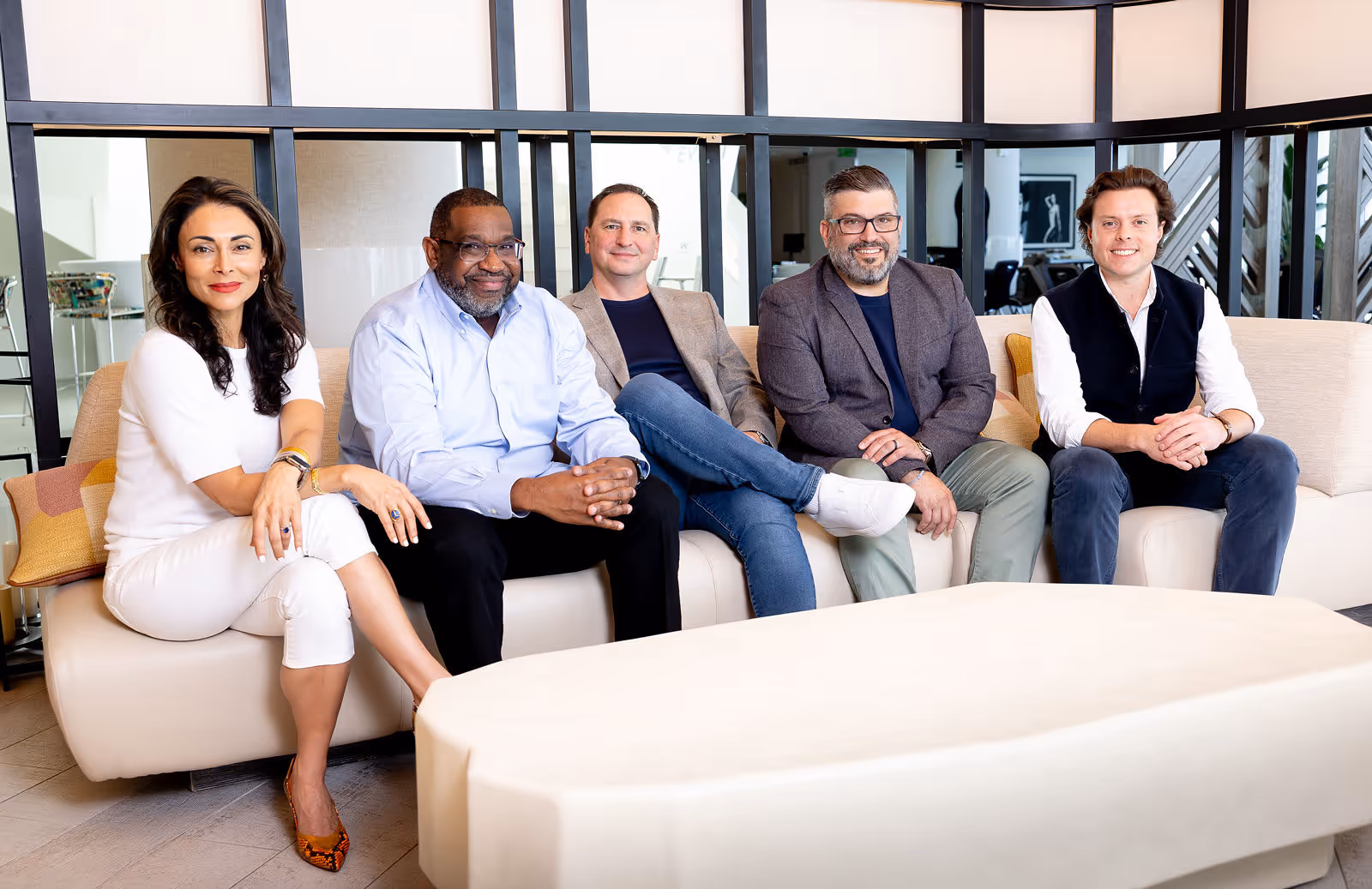 Five diverse professionals sitting on a beige couch in a modern office, smiling at the camera.