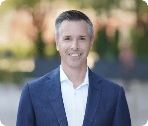 Smiling man with gray hair wearing a blue suit jacket and white shirt, with a blurred outdoor background.