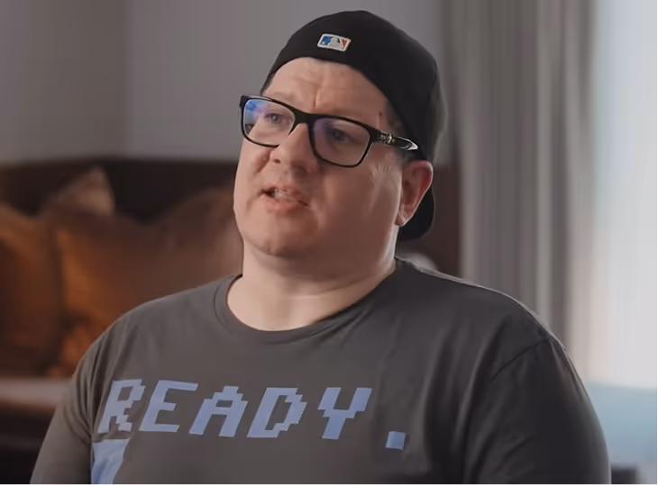 Man wearing glasses, a black backwards cap, and a dark t-shirt with the word 'READY.' printed on it, sitting indoors.