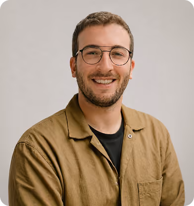 Smiling man with short brown hair and beard wearing glasses and a brown button-up shirt over a black t-shirt with a plain light gray background.