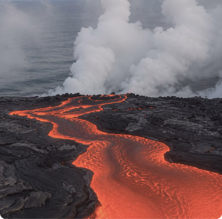 Flowing bright orange lava cutting through dark cooled volcanic rock with steam rising where lava meets the ocean.