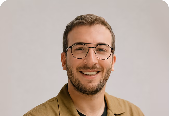 Smiling man with short brown hair, glasses, and light beard wearing a brown jacket over a black shirt, against a neutral background.