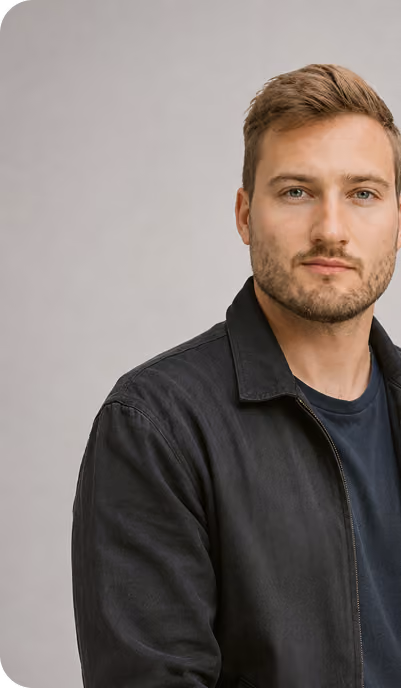 Portrait of a man with light brown hair and beard wearing a dark jacket over a navy shirt, standing against a plain gray background.
