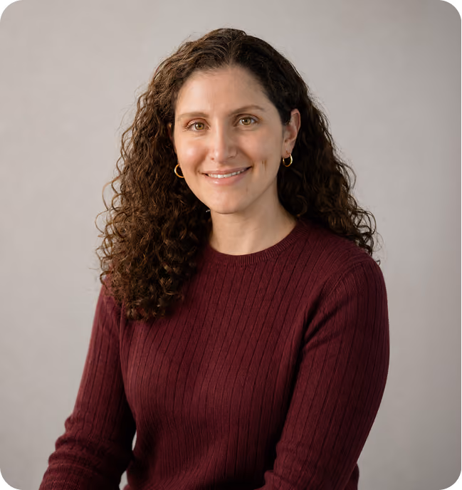 Woman with long curly brown hair, wearing a maroon ribbed sweater and gold hoop earrings, smiling against a plain light gray background.