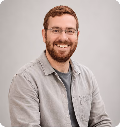Smiling man with glasses and a beard wearing a light gray buttoned shirt over a dark gray t-shirt against a plain light background.