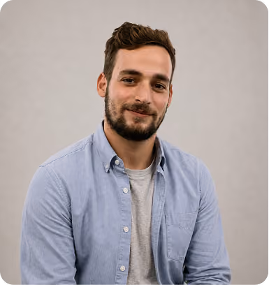 Portrait of a smiling man with short brown hair and beard wearing a light blue button-up shirt over a gray t-shirt against a plain light background.