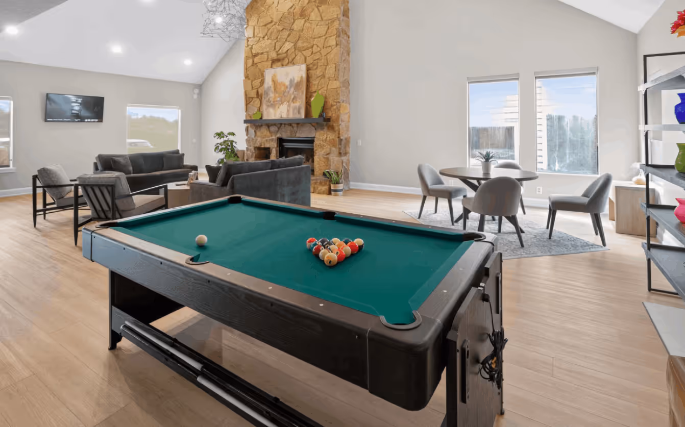 Modern game room with a green pool table, gray sofas, a stone fireplace, and a round table with four chairs near windows.