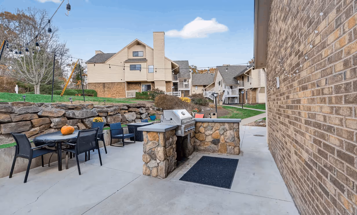 Outdoor patio area with stone-built barbecue grill, black chairs and tables, string lights, and multi-story beige residential buildings in the background.