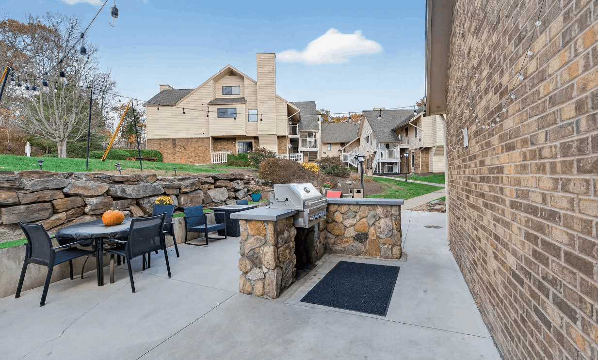 Outdoor patio area with stone-built barbecue grill, black chairs and tables, string lights, and multi-story beige residential buildings in the background.