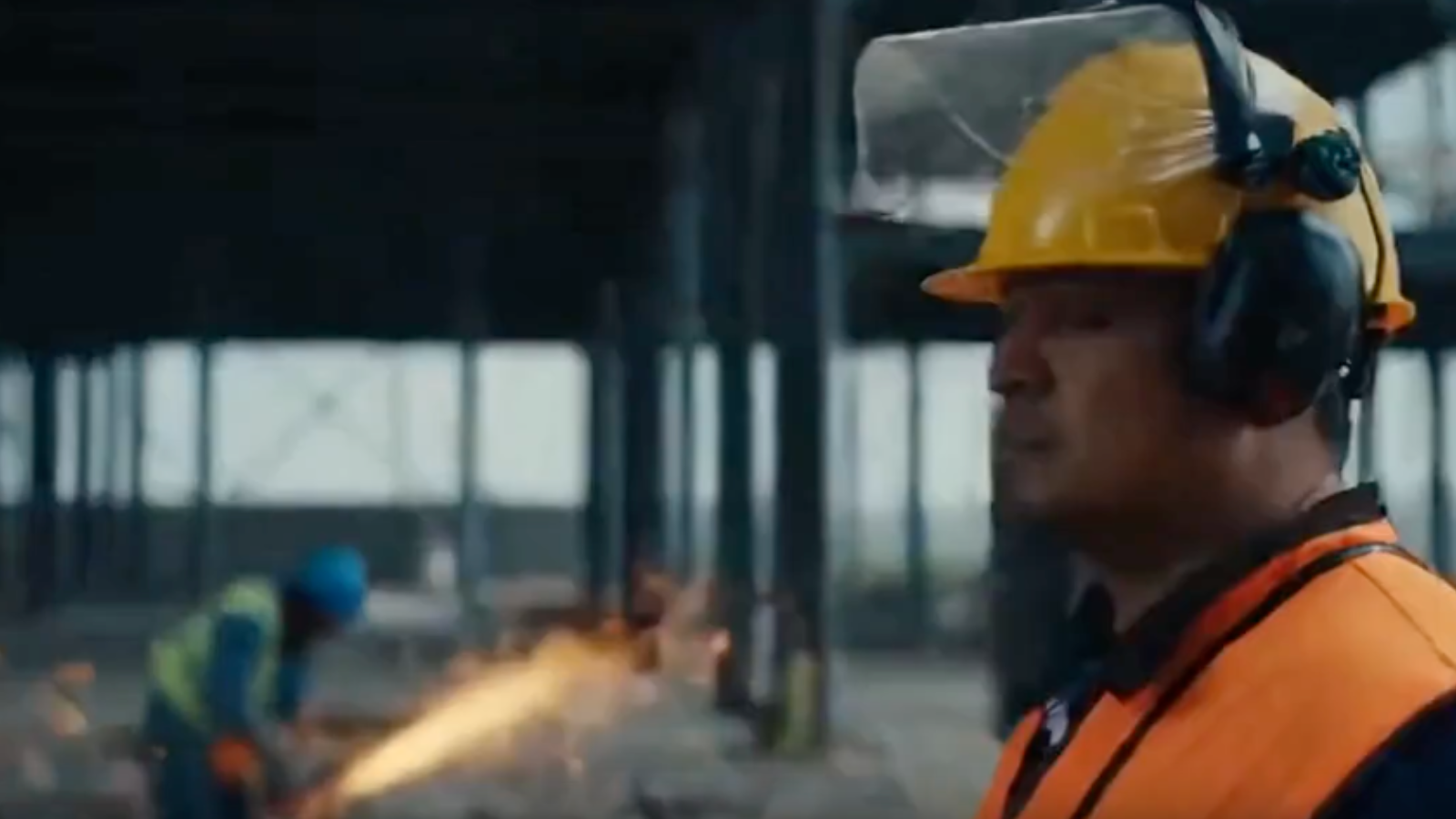 Construction worker wearing a yellow hard hat, orange safety vest, ear protection, and face shield on a building site with another worker using a welding torch in the background.