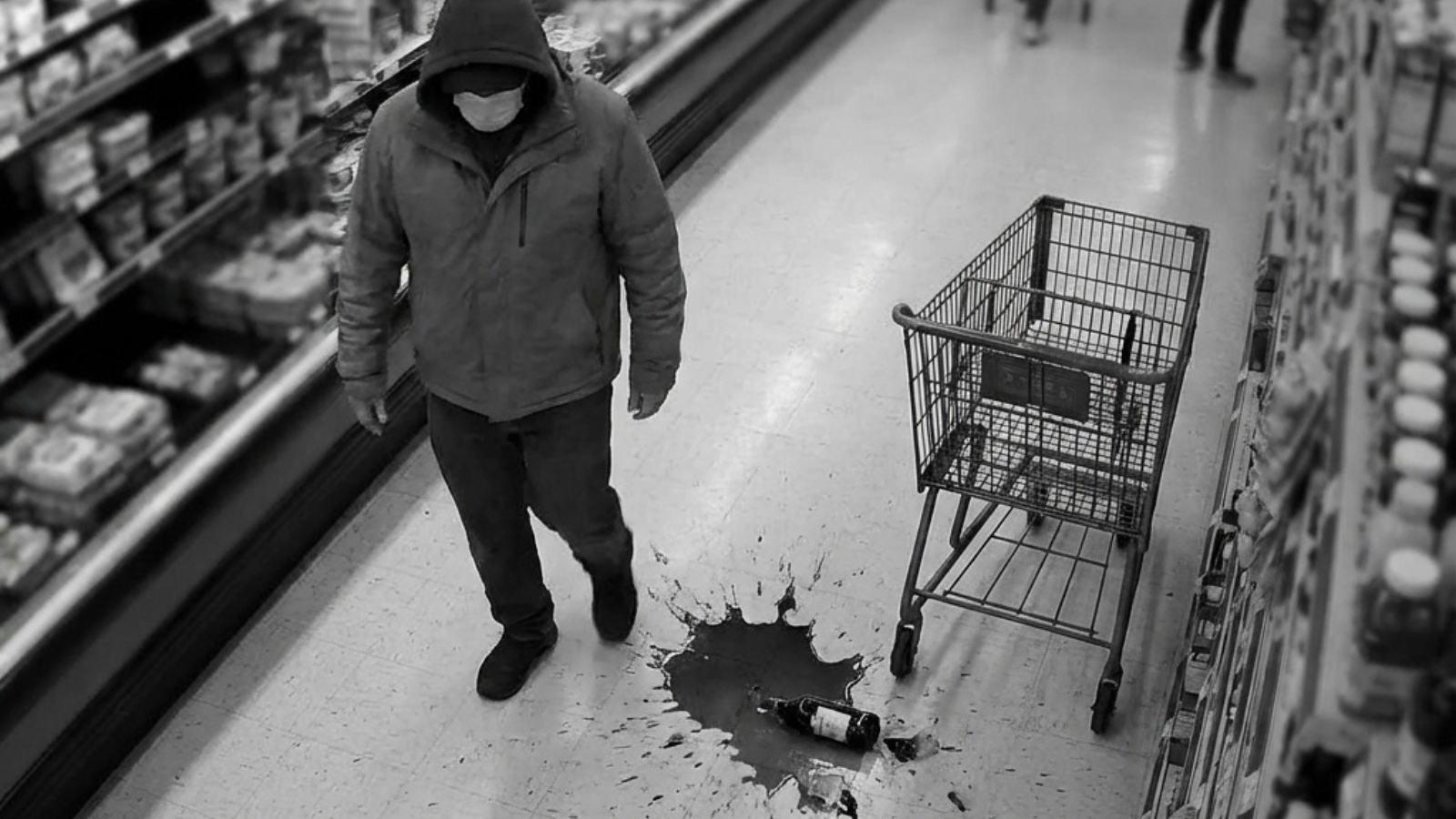 Person wearing a hood and face mask walking by a spilled bottle and liquid on a supermarket floor near an empty shopping cart.