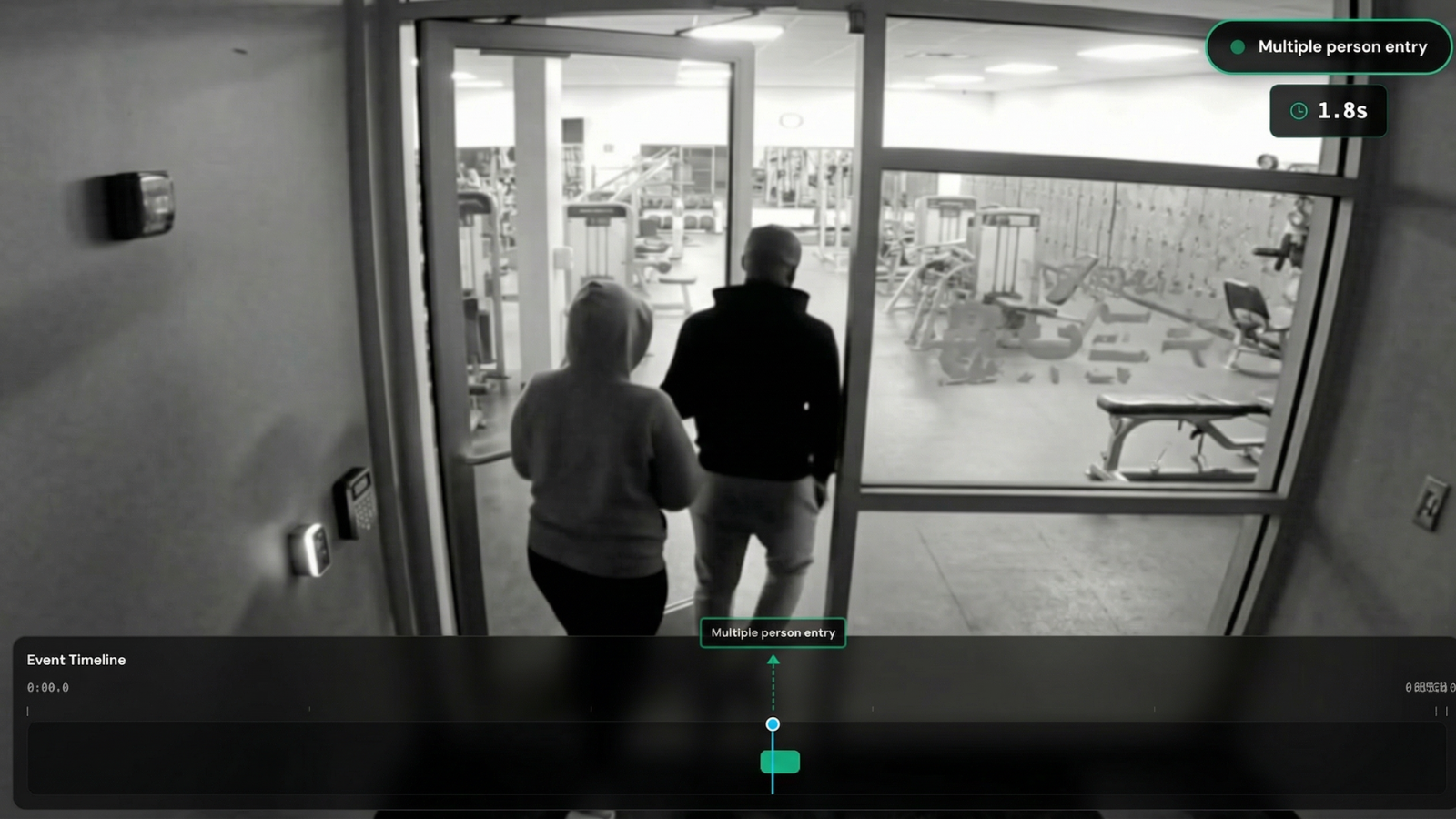 Two people entering a gym through a glass door, viewed from behind in a black and white surveillance image.