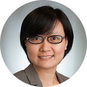 Portrait of an Asian woman with short black hair, wearing glasses and a brown blazer, smiling against a gray background.