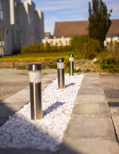 Drei Edelstahl-Gartenleuchten auf einem Bett aus weißen Kieselsteinen entlang eines gepflasterten Wegs.