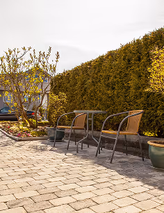 Zwei leere Gartenstühle mit kleinem Tisch auf gepflastertem Hof neben grüner Hecke und Blumentöpfen.