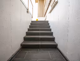 Graue Treppe mit Wandlampen an beiden Seiten in einem hell beleuchteten Korridor.