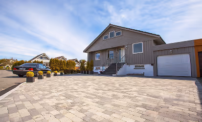 Moderner grauer Bungalow mit großer gepflasterter Einfahrt und geparktem Auto unter bewölktem Himmel.