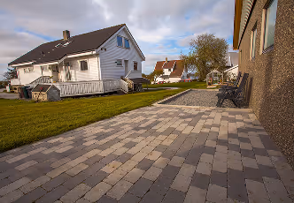 Wohnhaus mit weißer Fassade, gepflasterter Terrasse und grünem Rasen unter bewölktem Himmel.