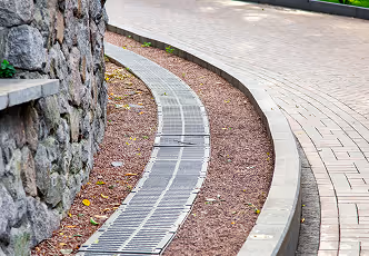 Gepflasterter Gehweg mit angrenzender Steingrundmauer und Bodenentwässerungsrinnen entlang einer gebogenen Kante.
