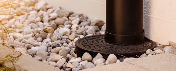 Schwarzes Fallrohr mit runder Gullyabdeckung umgeben von grauen Kieseln neben einem hellen Wandabschnitt.