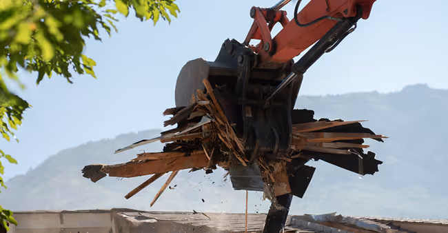 Baumaschine mit rot-orangenem Arm hebt zerbrochenes Holz vor einem Bergpanorama.