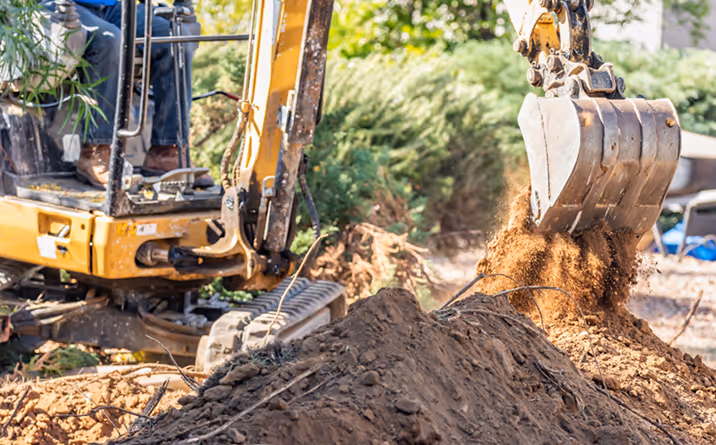 Ein Bagger hebt Erde von einem großen Erdhügel auf einer Baustelle bei Tageslicht.