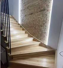 Moderner Holztreppenaufgang mit indirekter Beleuchtung an einer freiliegenden Backsteinwand.