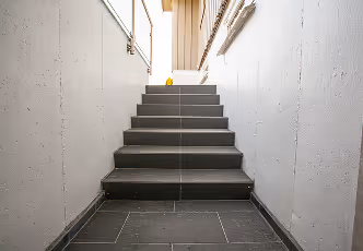 Innenraum mit grauen Stufen und weißen Wänden, steiler Treppenaufgang mit natürlichem Licht von oben.