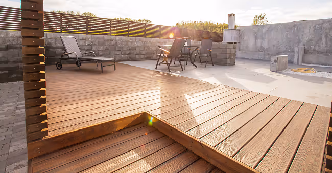 Terrasse mit Holzbelag, Sonnenlicht, zwei Liegestühlen und einem kleinen Tisch vor einer Steinmauer.