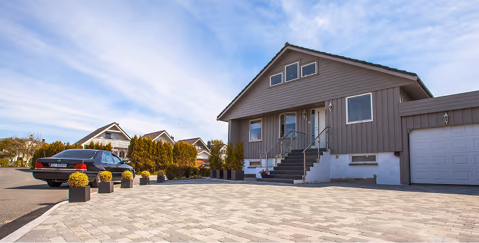 Ein modernes graues Einfamilienhaus mit einer gepflasterten Einfahrt und einem schwarzen Auto, umgeben von Büschen und anderen Häusern bei klarem Himmel.