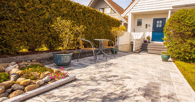 Terrasse mit gepflastertem Boden, zwei Stühlen und kleinem Tisch vor einem weißen Haus mit blauer Tür und grünen Pflanzen in Töpfen.